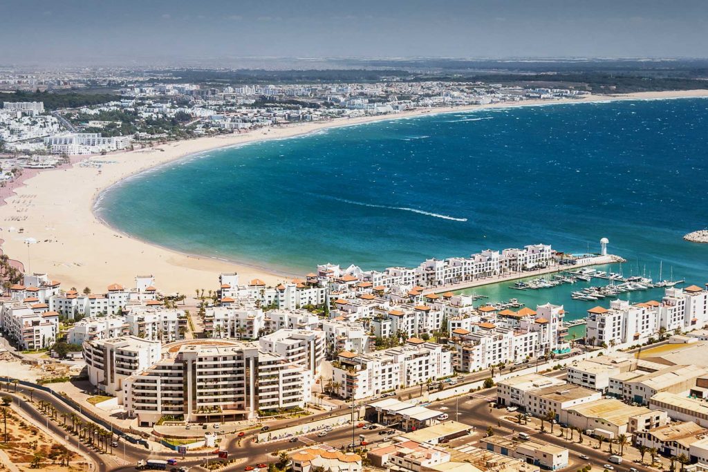 A high-angle aerial view of Agadir, Morocco, showcasing a massive, crescent-shaped sandy beach bordering a vibrant turquoise and deep blue ocean. In the foreground, a modern marina is filled with moored yachts and boats, flanked by uniform white-washed luxury apartments and resort complexes with terracotta roofs. A coastal road curves along the bottom of the frame, and the sprawling city stretch extends far into the hazy background along the coastline under a clear sky.
