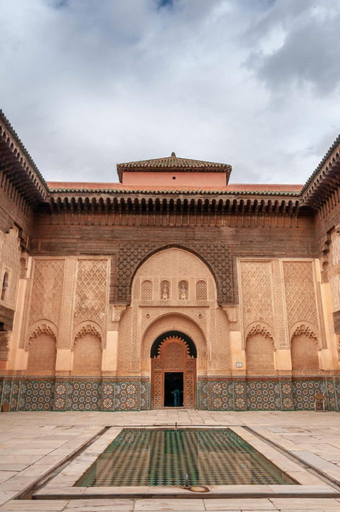 A symmetric, eye-level shot of the inner courtyard of a historic Moroccan madrasa or palace. The courtyard features a long, rectangular reflection pool lined with green tiles in the foreground. In the center backdrop, a beautifully carved arched wooden doorway leads into a dim interior room. The surrounding walls are heavily decorated with intricate geometric stucco carvings, a lower band of colorful mosaic tiles, and dark, detailed cedar wood corbels supporting a green tiled roof under a cloudy sky.