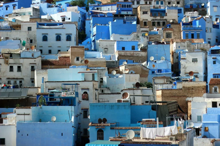 A densely packed hillside view of Chefchaouen, Morocco, showcasing traditional buildings painted in various shades of blue and white. The structures are tightly clustered together, featuring flat roofs dotted with satellite dishes, small windows, and terracotta tiled awnings. In the lower foreground, a white clothesline with laundry hanging out to dry is visible on a rooftop.