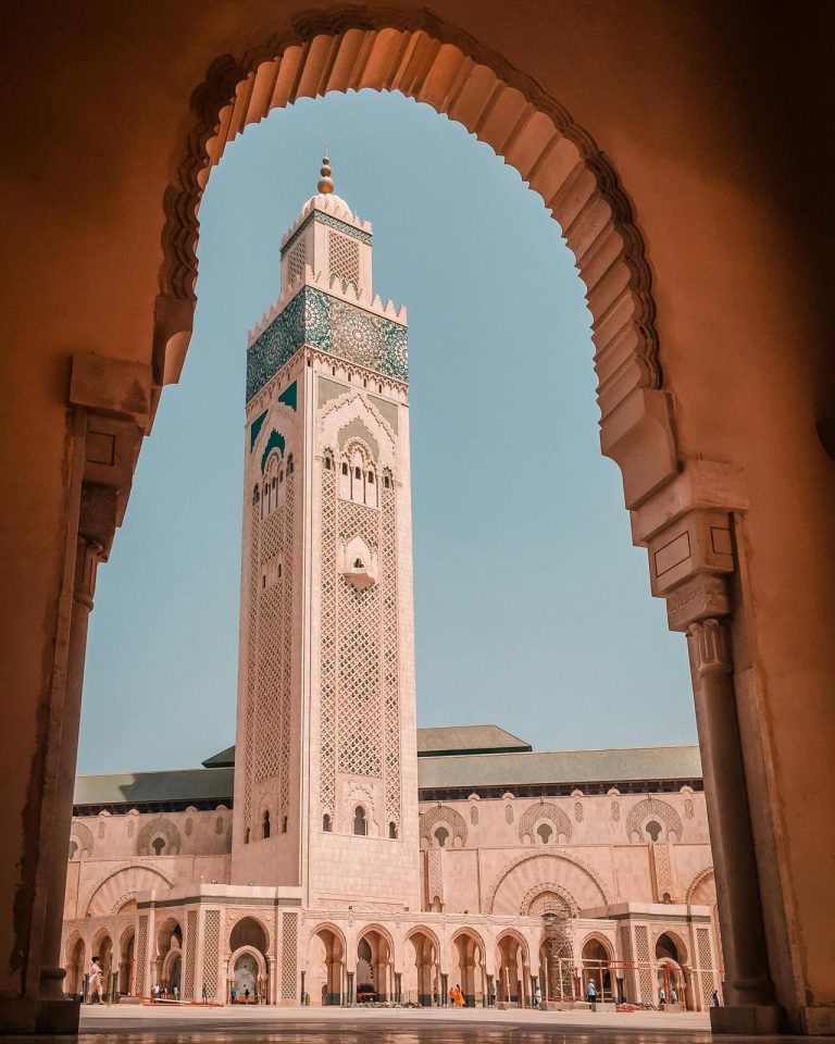 A low-angle shot of the towering minaret of the Hassan II Mosque in Casablanca, framed perfectly by a large, ornate stone archway in the foreground. The massive cream-colored minaret features intricate geometric carvings and a prominent band of green and blue mosaic tilework near its top. The main mosque building extends out below with a long arcade of arches under a pale, clear blue sky.