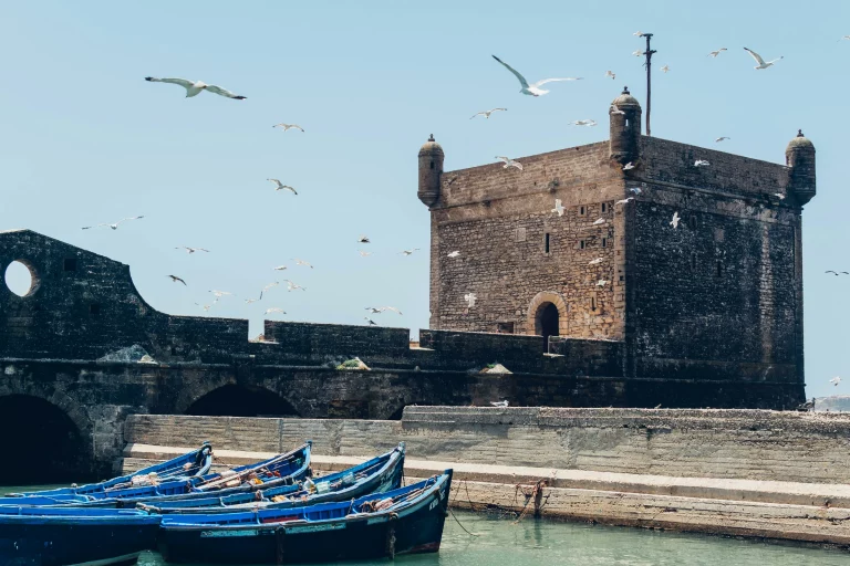 A sunny, wide shot of the stone Sqala du Port fortress in Essaouira, Morocco. In the foreground, several traditional bright blue wooden fishing boats sit in the calm green harbor water next to a concrete pier. The massive, weathered brown stone bastion features circular corner watchtowers and arched openings. A flock of seagulls flies across the clear, bright blue sky surrounding the historical fortifications.