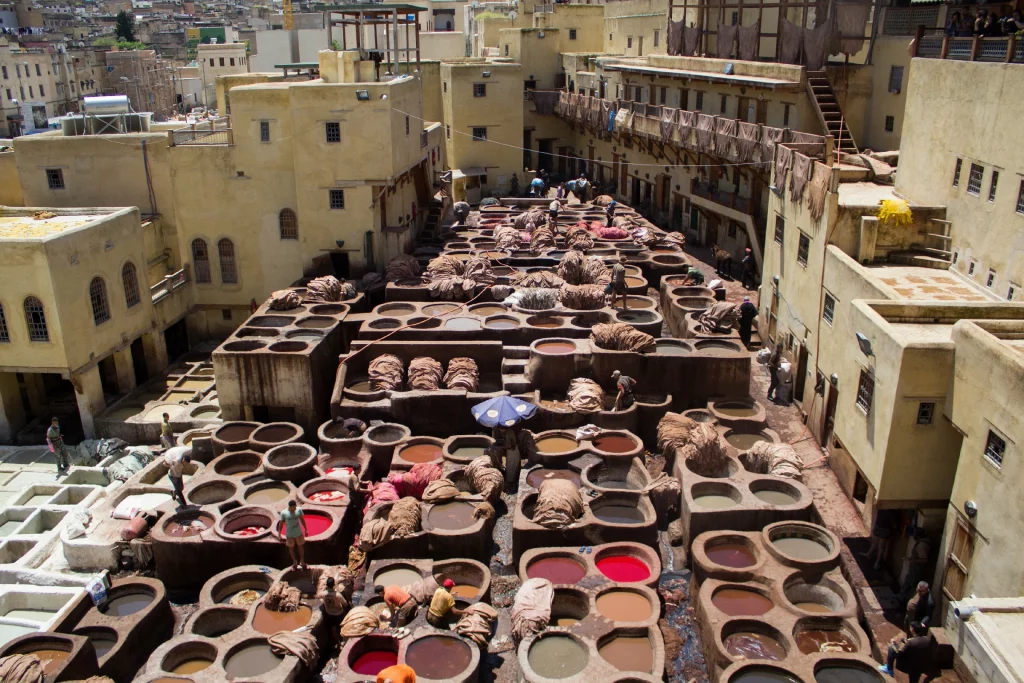 A high-angle, wide shot of the historic Chouara Tannery in Fes, Morocco. The open-air courtyard is packed with numerous round, stone dye vats filled with various colored liquids like red, brown, and white. Several tanners are working among the pits, processing and treating bundles of animal hides. The tannery is entirely enclosed by multi-story, sand-colored traditional buildings with balconies where dyed hides hang to dry.