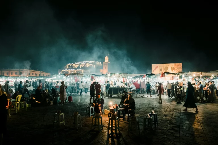 A wide, atmospheric night photograph of the vibrant Jemaa el-Fnaa public square in Marrakech. The background features rows of tightly packed, brightly lit open-air food stalls with white lights, under a hazy layer of smoke rising from outdoor grills into the dark night sky. Numerous locals and tourists walk among the stalls or congregate in groups. In the foreground, empty plastic and wooden stools are scattered across the dark, tiled stone ground, where a few vendors sit illuminated by small lanterns. In the distance, illuminated traditional buildings and a small minaret tower rise against the dark horizon.