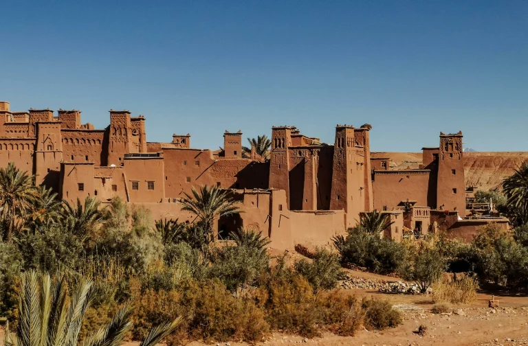 A wide, sunny view of a historic clay-and-straw fortified village, or Ksar, in Morocco, resembling Ait Benhaddou. The sprawling multi-story complex features traditional earthen red-brown mud-brick architecture, prominent square watchtowers with defensive slot windows, and decorative crenellations along the rooflines. Lush green palm trees and desert shrubs populate the foreground under a completely clear, bright blue sky.