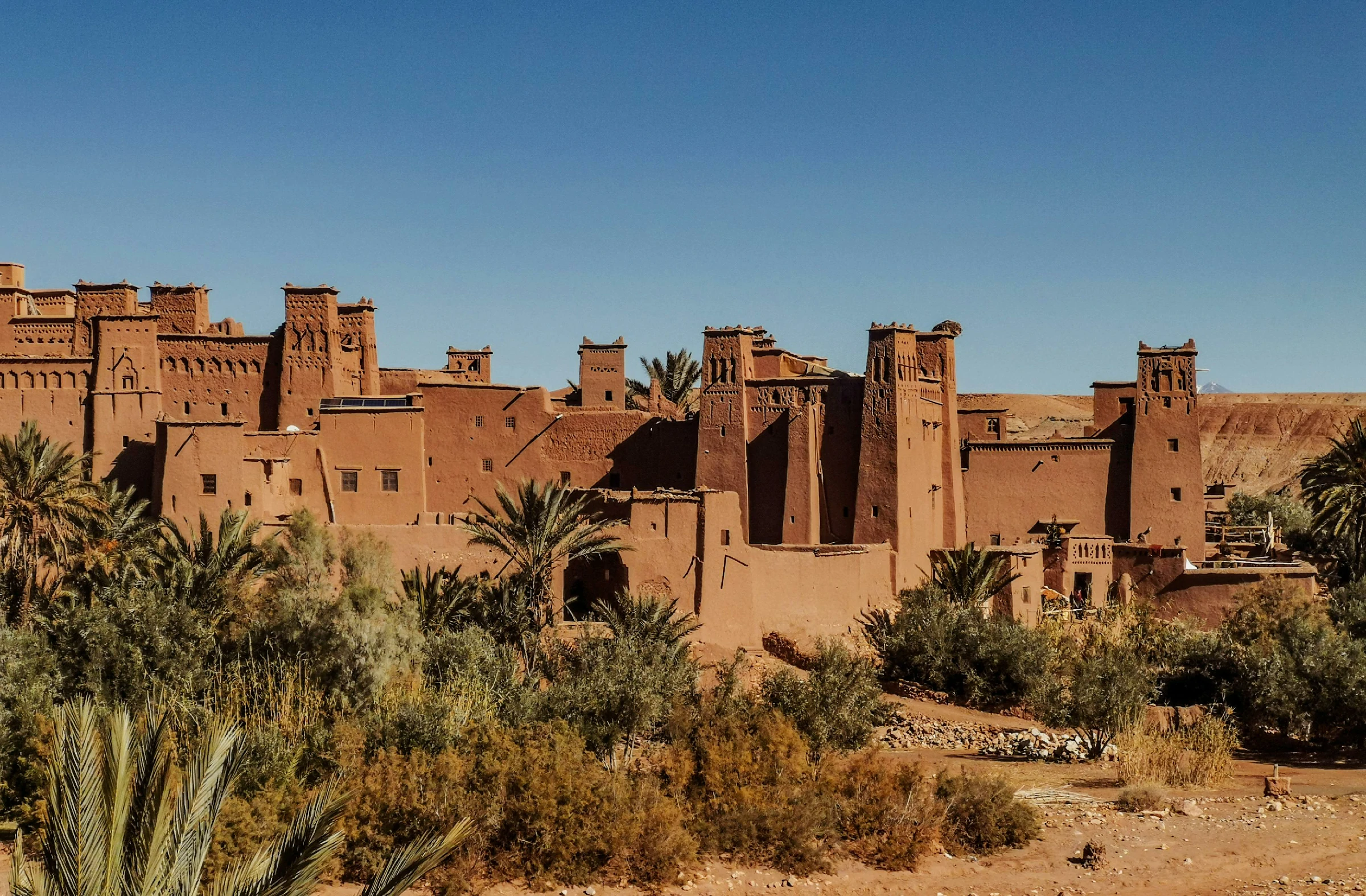 A wide, sunny view of a historic clay-and-straw fortified village, or Ksar, in Morocco, resembling Ait Benhaddou. The sprawling multi-story complex features traditional earthen red-brown mud-brick architecture, prominent square watchtowers with defensive slot windows, and decorative crenellations along the rooflines. Lush green palm trees and desert shrubs populate the foreground under a completely clear, bright blue sky.