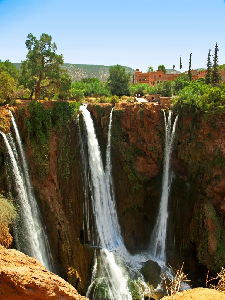 A high-angle, vertical photograph of a dramatic, multi-tiered waterfall cascading down a steep gorge of reddish-brown rock. Multiple distinct streams of white water plunge into the canyon below, with green moss and climbing vegetation clinging to the cliff faces. The upper edge of the falls is lined with rich green trees and shrubbery. In the upper right background, traditional terracotta-colored, mud-brick style buildings are nestled among the trees. Rolling green hills rise in the far distance under a bright, clear blue sky.