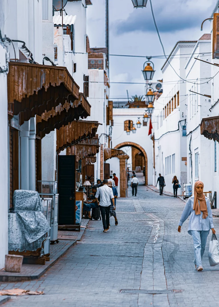 A bustling cobblestone street in the old medina of Rabat, Morocco. The street is lined with traditional whitewashed buildings featuring distinctive, ornate brown wooden awnings over storefronts. Several pedestrians are walking along the pathway, including a woman in the foreground wearing a hijab, light blue shirt, and jeans, and a man walking away in a white shirt. String lights with glass lanterns hang overhead, leading toward a stone archway in the distance under a cloudy sky.