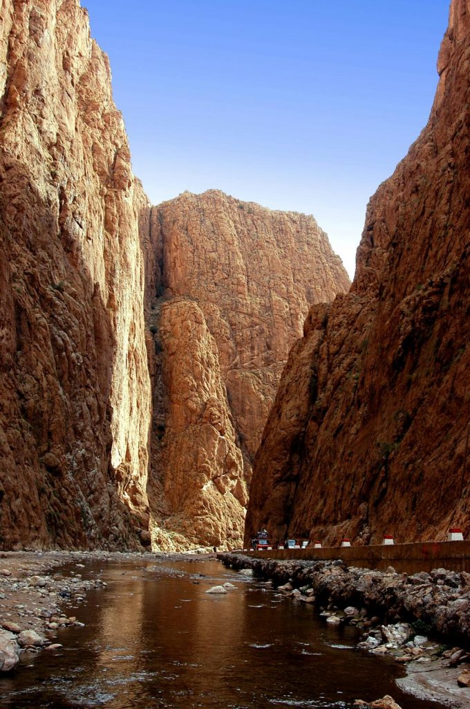 A high-angle, vertical landscape photograph looking through a massive, narrow canyon. Towering, sheer limestone cliffs in shades of orange and rugged brown rise steeply on both the left and right sides of the frame, completely dominating the view. At the bottom of the gorge, a shallow, dark river flows toward the foreground, reflecting the rocky walls. On the right bank, a narrow asphalt road with small, white-and-red concrete safety markers runs along the base of the cliff, with a few distant vehicles parked. A colossal rock formation stands prominently in the center background where the canyon curves, all set beneath a bright, clear blue sky.