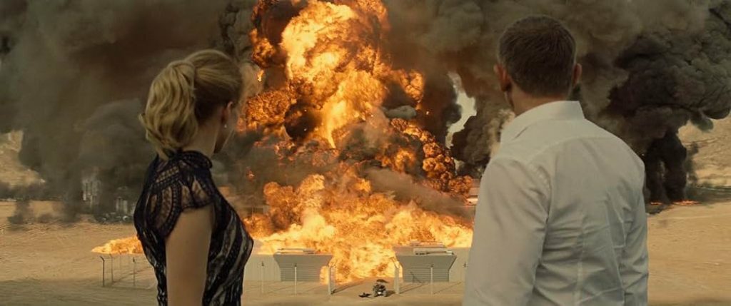 A cinematic wide shot taken from behind James Bond (Daniel Craig) and Madeleine Swann (Léa Seydoux) as they stand in a desert landscape looking toward a massive, fiery explosion. James Bond is on the right, wearing a crisp white dress shirt, and Madeleine is on the left with her blonde hair in a ponytail, wearing a dark lace dress. Centered in front of them, a colossal fireball of orange flame and thick, billowing black and grey smoke erupts violently over a fenced facility, filling the majority of the background.