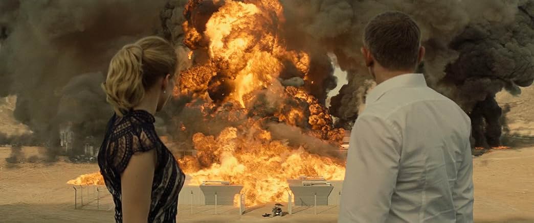 A cinematic wide shot taken from behind James Bond (Daniel Craig) and Madeleine Swann (Léa Seydoux) as they stand in a desert landscape looking toward a massive, fiery explosion. James Bond is on the right, wearing a crisp white dress shirt, and Madeleine is on the left with her blonde hair in a ponytail, wearing a dark lace dress. Centered in front of them, a colossal fireball of orange flame and thick, billowing black and grey smoke erupts violently over a fenced facility, filling the majority of the background.