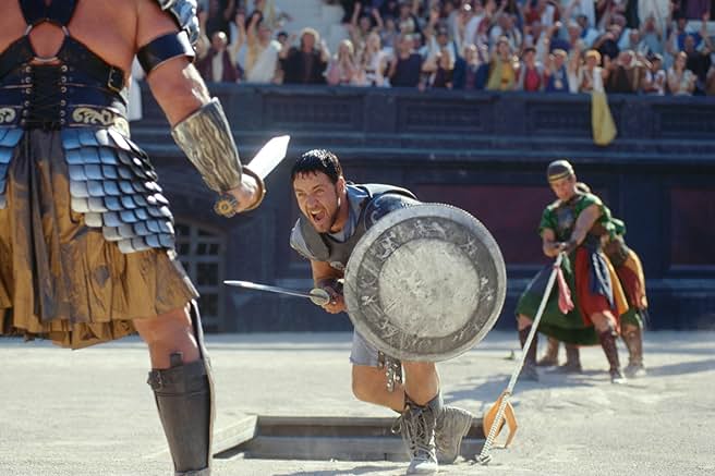 A dynamic mid-shot from the movie Gladiator. In the center, Maximus (Russell Crowe) charges forward with an intense, shouting expression, wielding a short sword in his right hand and holding a round, metal shield with intricate engravings in his left. In the foreground on the left, the armored back and shoulder of a large gladiator opponent are visible, holding a sword pointed toward Maximus. In the background to the right, another arena fighter stands pulling a taut rope, while a crowded stadium of spectators cheers from the sunlit grandstands behind a dark arena wall.